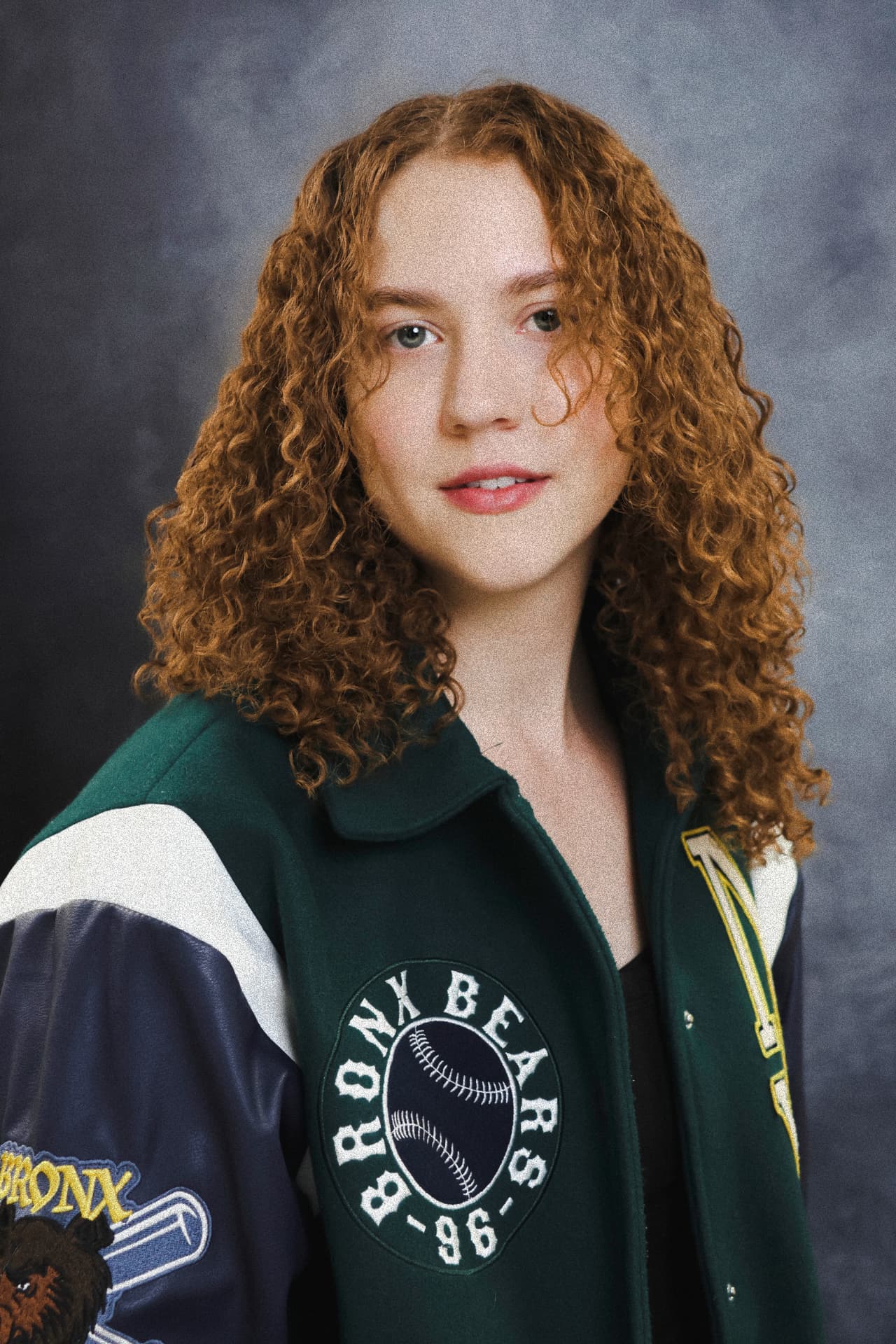 Actor portrait of a person with curly auburn hair in a varsity jacket, photographed against a grey backdrop at the Ceeb Moz studio in King's Cross, London.
